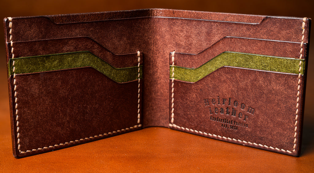 Brown leather wallet with green accents on a wooden surface, featuring the brand 'Heirloom Leather'.