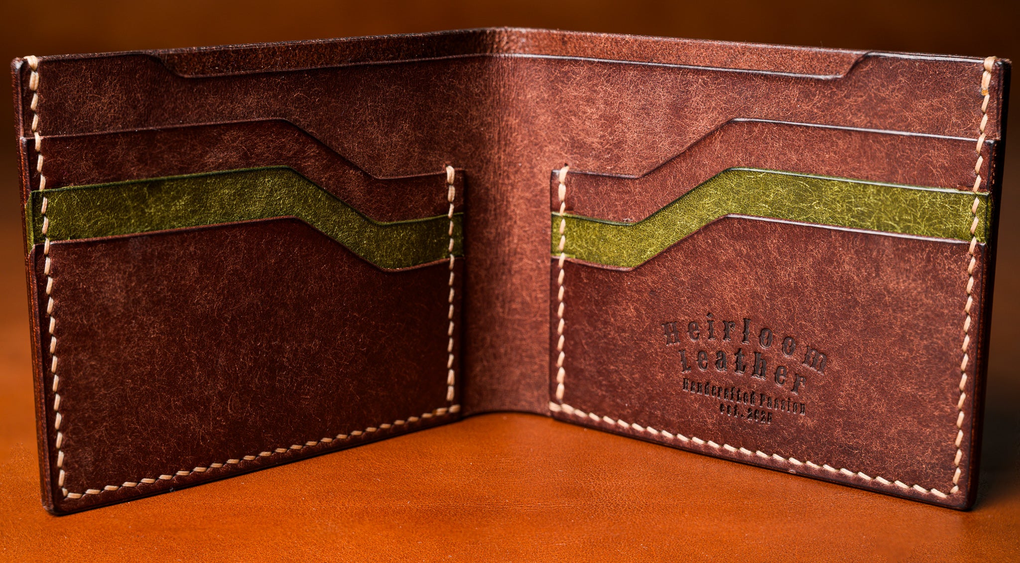 Brown leather wallet with green accents on a wooden surface, featuring the brand 'Heirloom Leather'.